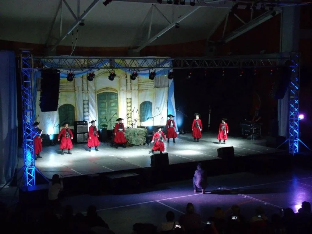 Esperando el 9 de Julio: la vigilia patriótica que Ushuaia vive a puro folclore Grupo de bailarines con vestuario rojo tradicional en escenario iluminado durante espectáculo nocturno, Ushuaia